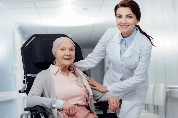 Beautiful happy female doctor near senior woman in kerchief with cancer sitting in wheelchair in hospital — Stock Photo