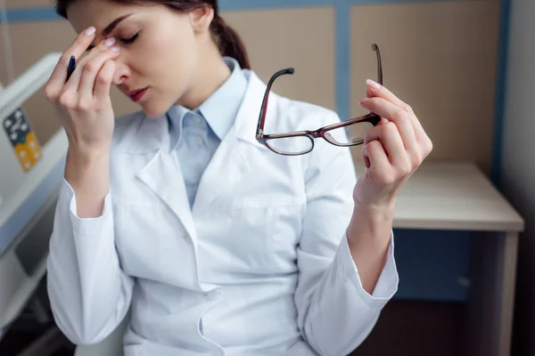 Cansada doctora sosteniendo gafas, tocando la frente y teniendo dolor de cabeza en el hospital - foto de stock