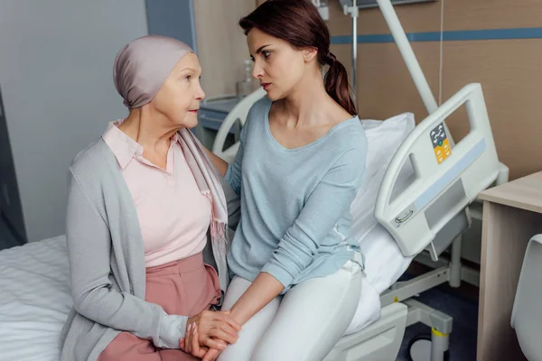 Figlia preoccupata seduta vicino alla madre anziana con il cancro sul letto e che si tiene per mano in ospedale — Foto stock