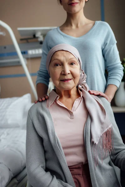 Femme âgée souriante en mouchoir regardant la caméra avec sa fille en arrière-plan à l'hôpital — Photo de stock
