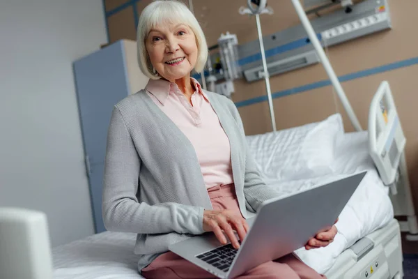Усміхнена старша жінка сидить на ліжку і використовує ноутбук у лікарні — стокове фото