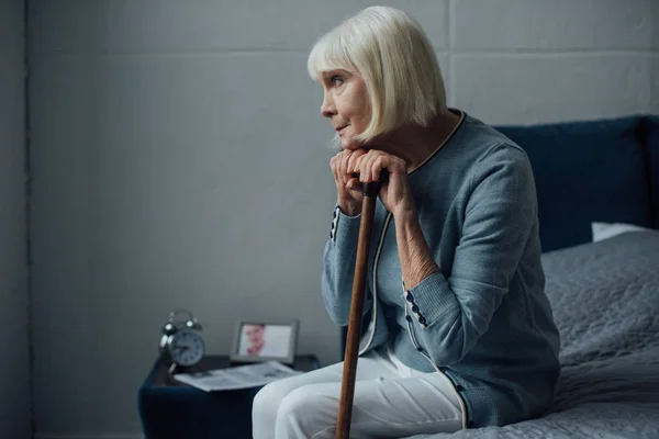 Donna anziana sola con le mani sul bastone da passeggio seduto sul letto a casa — Foto stock
