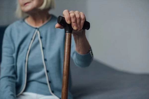 Partial view of senior woman sitting on bed and holding walking stick at home — Stock Photo