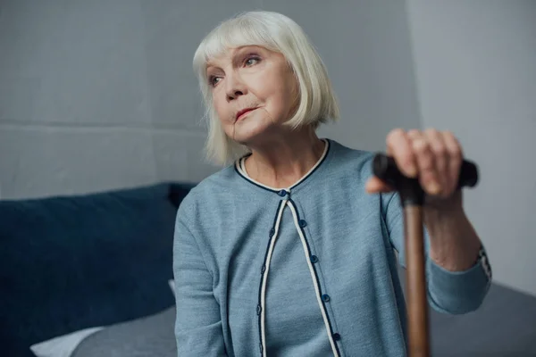 Lonely senior woman sitting on bed with walking stick at home — Stock Photo