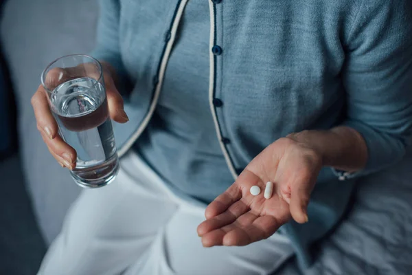 Vista cortada da mulher sênior sentado e segurando pílulas com vidro de água em casa — Fotografia de Stock