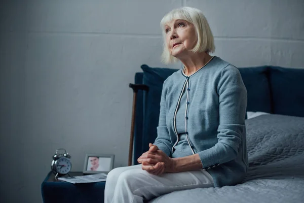 Chère femme âgée assise sur le lit avec les mains pliées à la maison et regardant loin — Photo de stock