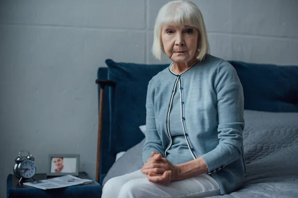 Mujer mayor mirando a la cámara y sentada en la cama con las manos dobladas en casa - foto de stock