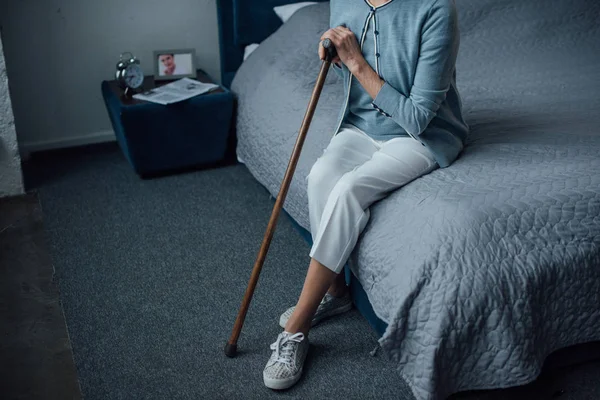 Vue partielle de la femme âgée assise sur le lit avec bâton de marche à la maison — Photo de stock