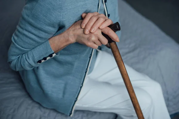 Vista recortada de la mujer mayor sentada en la cama con bastón en casa - foto de stock