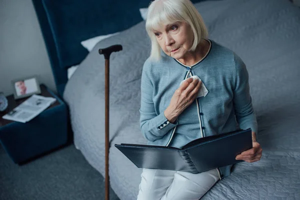 Traurige Seniorin sitzt auf Bett und hält Fotoalbum mit Taschentuch zu Hause — Stockfoto