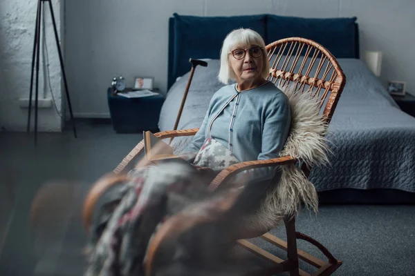 Sad senior woman in glasses sitting in wicker rocking chair at home — Stock Photo