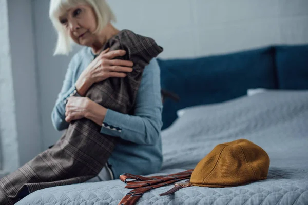 Sad senior woman sitting on bed and holding jacket at home — Stock Photo