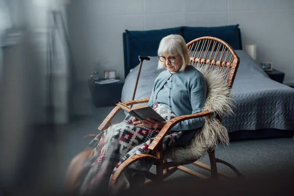 Старша жінка сидить у плетеному кріслі і читає книгу в спальні — стокове фото