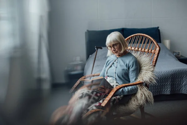 Femme âgée assise dans un fauteuil berçant en osier et un livre de lecture à la maison — Photo de stock