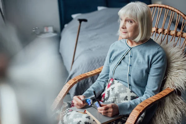 Donna anziana pensierosa seduta in sedia a dondolo di vimini con libro a casa — Foto stock