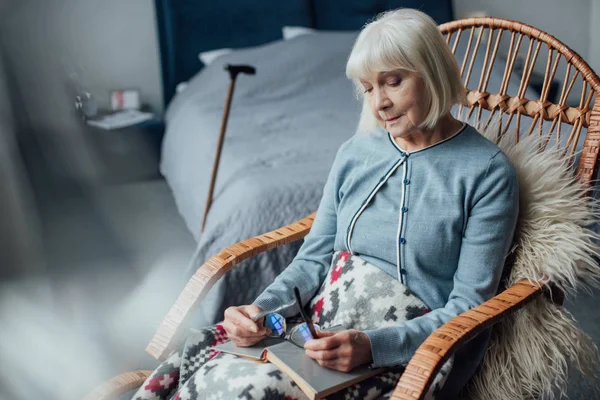 Pensativa mujer mayor sentada en mecedora de mimbre con libro en casa - foto de stock