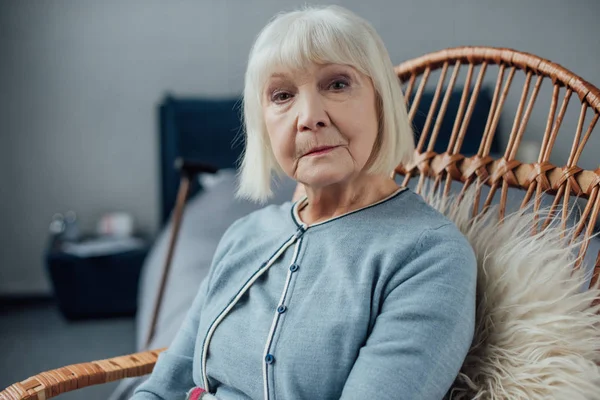 Seria donna anziana seduta in sedia a dondolo di vimini a casa e guardando la fotocamera — Foto stock