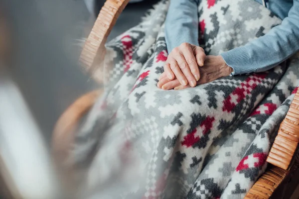 Vista ritagliata della donna anziana seduta in vimini sedia a dondolo con coperta e mani piegate a casa — Foto stock