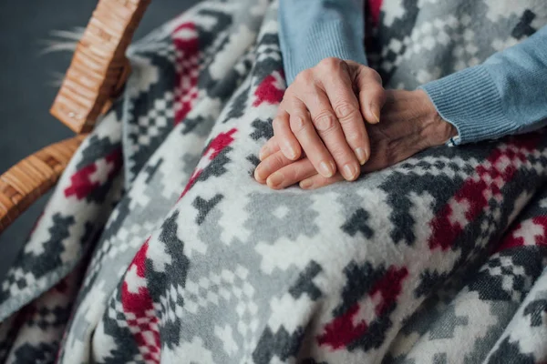 Vista parziale della donna anziana seduta in sedia a dondolo in vimini con coperta e mani piegate a casa — Foto stock