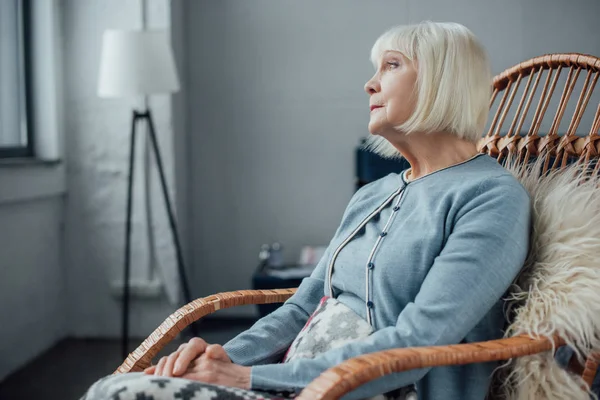 Femme âgée assise dans un fauteuil berçant en osier à la maison et regardant loin — Photo de stock