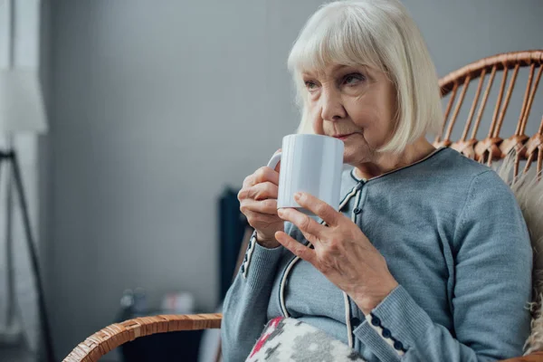 Donna anziana seduta in vimini sedia a dondolo e bere caffè a casa — Foto stock