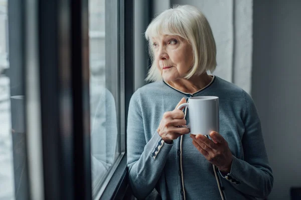 Donna anziana seduta vicino alla finestra e bere caffè a casa — Foto stock