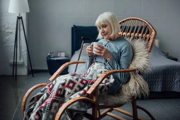 Senior femme assise dans une chaise berçante en osier avec couverture et tenant une tasse de café à la maison — Photo de stock