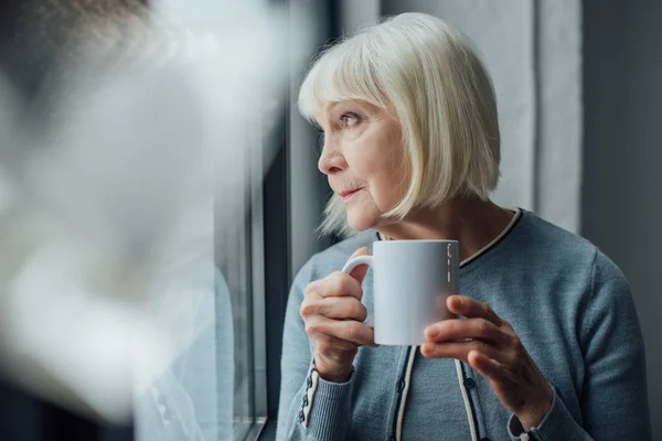 Femme âgée réfléchie tenant une tasse de café à la maison et regardant loin — Photo de stock