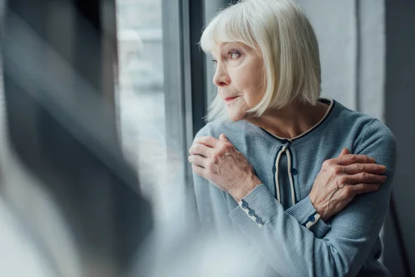 Nachdenkliche Seniorin mit gekreuzten Händen schaut durch Fenster nach Hause — Stockfoto