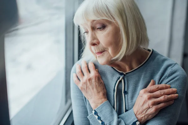 Triste donna anziana con le mani incrociate vicino alla finestra a casa — Foto stock