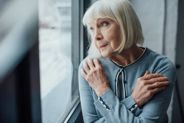 Nachdenkliche Seniorin mit verschränkten Händen schaut durch Fenster nach Hause — Stockfoto