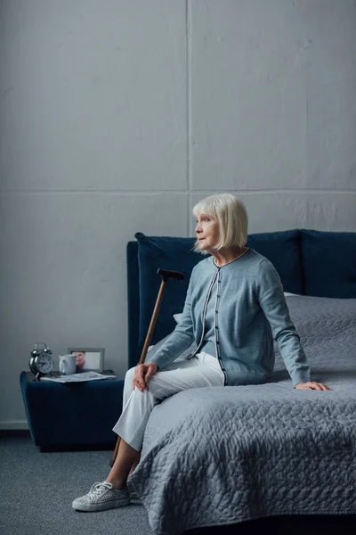 Задумчивая старшая женщина сидит на кровати с тростью дома — стоковое фото
