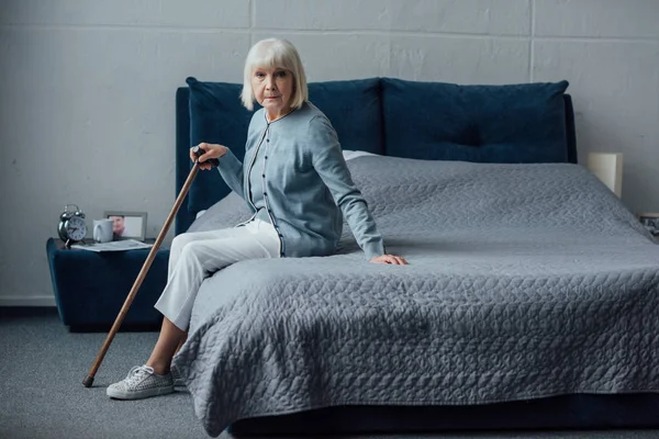 Seniorin sitzt zu Hause mit Gehstock im Bett und blickt in Kamera — Stockfoto