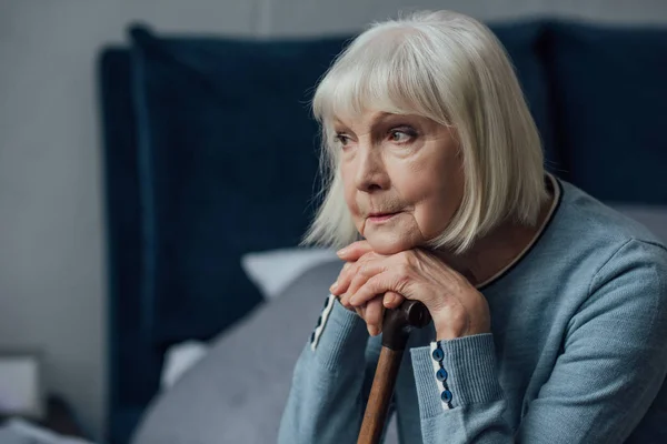 Femme âgée réfléchie avec les mains sur bâton de marche assis sur le lit à la maison — Photo de stock