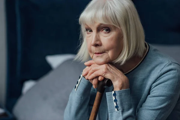 Nachdenkliche Seniorin mit den Händen am Gehstock sitzt zu Hause auf dem Bett und blickt in die Kamera — Stockfoto