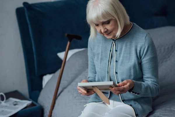 Senior woman sitting on bed and looking at photo frame at home — Stock Photo