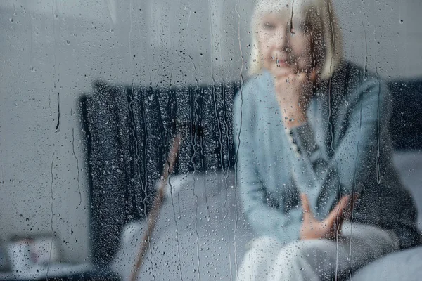 Самотня старша жінка сидить на ліжку і підборіддя з рукою вдома через вікно з краплями дощу — стокове фото