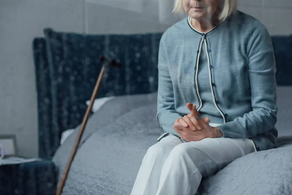 Visão parcial da mulher idosa sentada na cama com as mãos dobradas em casa — Fotografia de Stock