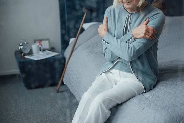 Abgeschnittene Ansicht einer einsamen Seniorin, die zu Hause mit gekreuzten Händen auf dem Bett sitzt — Stockfoto