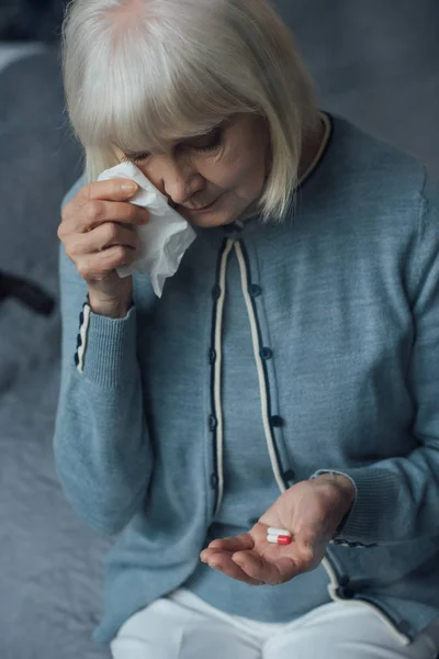 Aufgebrachte Seniorin sitzt auf Bett, hält Tabletten und wischt sich zu Hause Tränen mit Taschentuch ab — Stockfoto