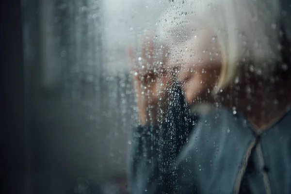 Blurred portrait of lonely senior woman at home through window with raindrops — Stock Photo