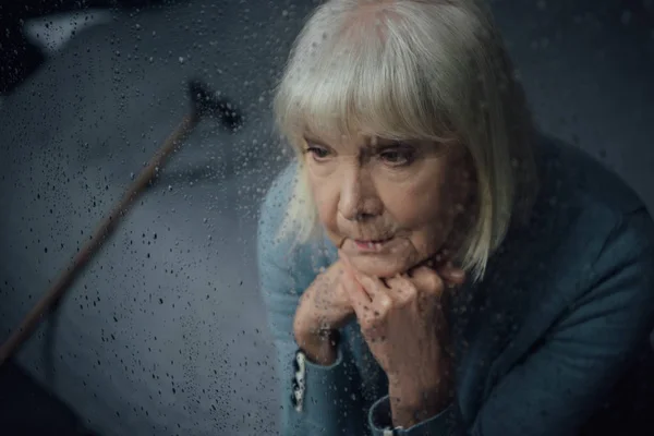 Depressed senior woman sitting and propping chin with hands at home through window with raindrops — Stock Photo