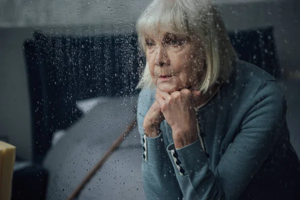 Upset senior woman sitting and propping chin with hands at home through window with raindrops — Stock Photo
