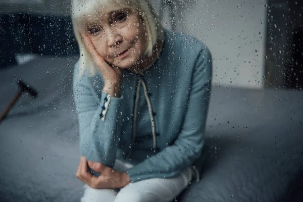 Самотня старша жінка сидить і дивиться на камеру через вікно з краплями дощу — стокове фото