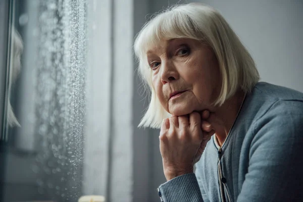 Femme âgée réfléchie avec les cheveux gris appui menton avec la main et en regardant la caméra à la maison — Photo de stock