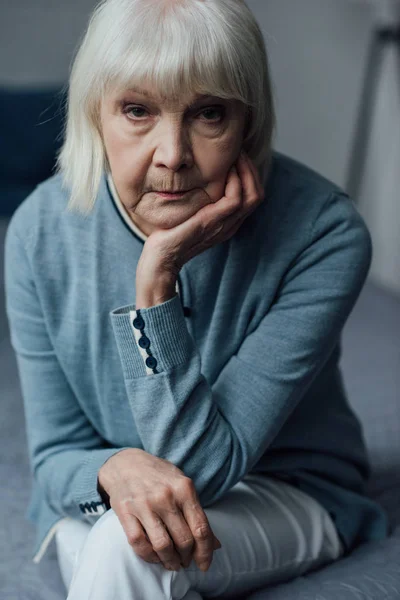Mujer mayor disgustada con el pelo gris apoyando la barbilla con la mano y mirando a la cámara en casa - foto de stock