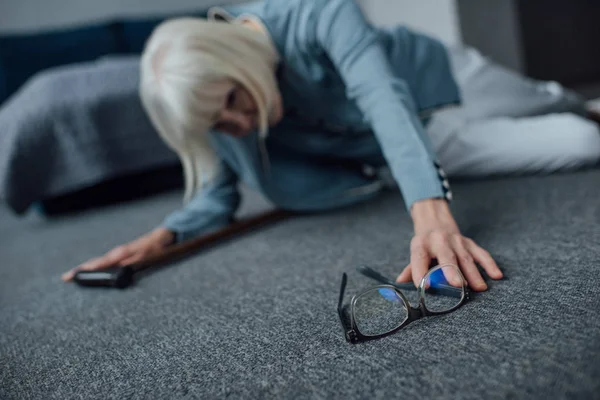 Femme âgée solitaire allongée sur le sol, se sentant mal et cherchant des lunettes à la maison — Photo de stock