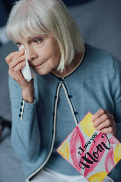 Senior woman holding card with 'i love you mom' lettering, crying and wiping tears with handkerchief at home — Stock Photo