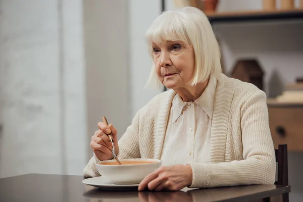 Расстроена старшая женщина сидит за столом и ест сливочный суп дома — стоковое фото