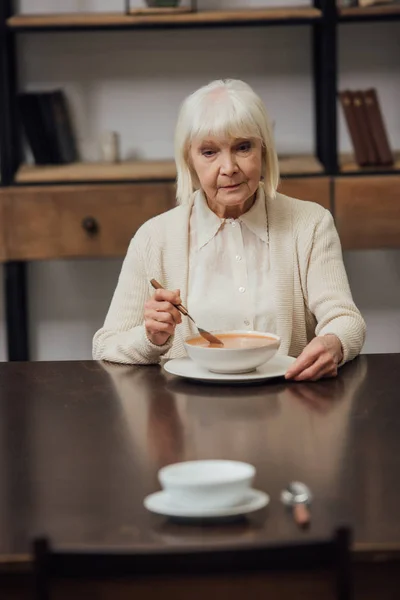 Selective focus of sad senior woman eating at table with bowl and spoon on foreground — Stock Photo